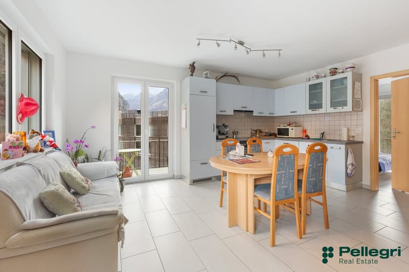 Open-plan living room and kitchen, with large windows and sliding glass doors leading to a balcony. The kitchen has white cabinets and appliances, and the dining area has a wooden table with orange chairs. The living room has a gray sofa and decorative it
