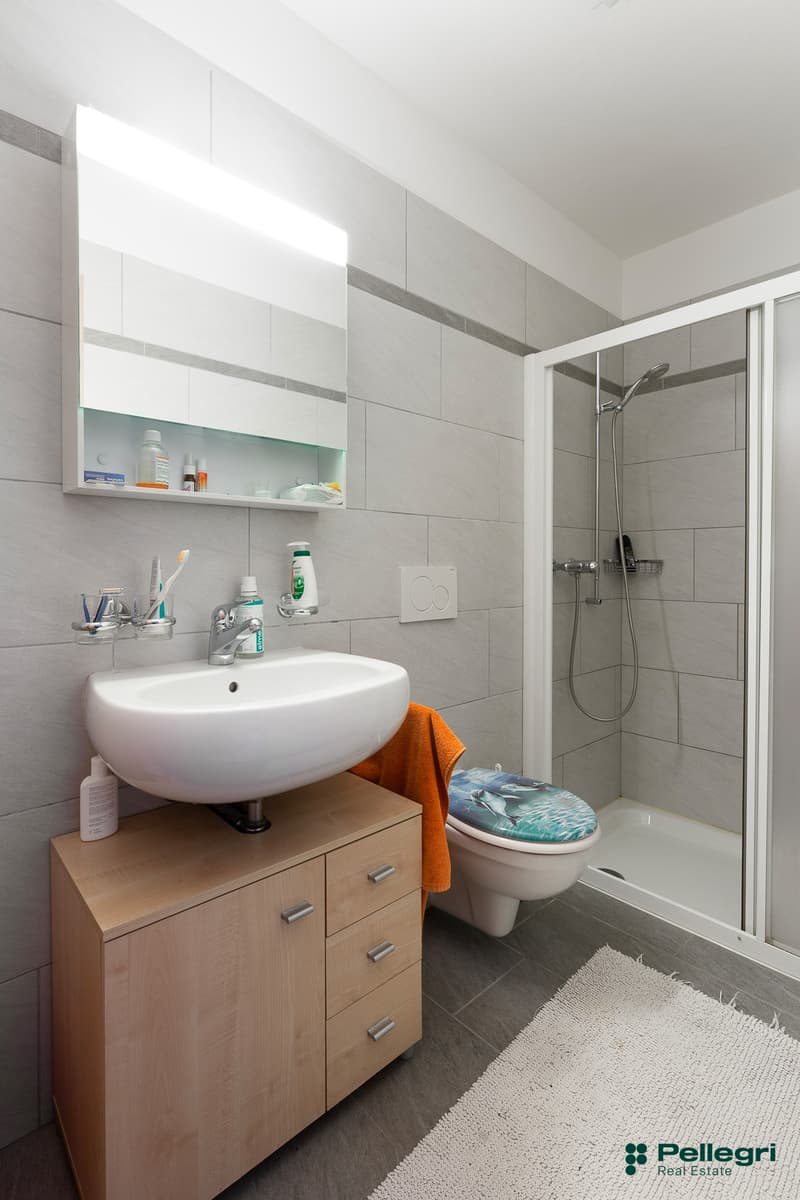 Modern bathroom with gray tile walls, white sink, wooden vanity cabinet, toilet, and shower enclosure