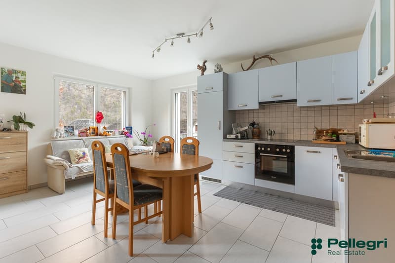 The image shows an open-plan kitchen and dining area. The kitchen features white cabinets, a tiled backsplash, and a built-in oven and stovetop. The dining area has a round wooden table with four chairs. The room has large windows that provide natural lig