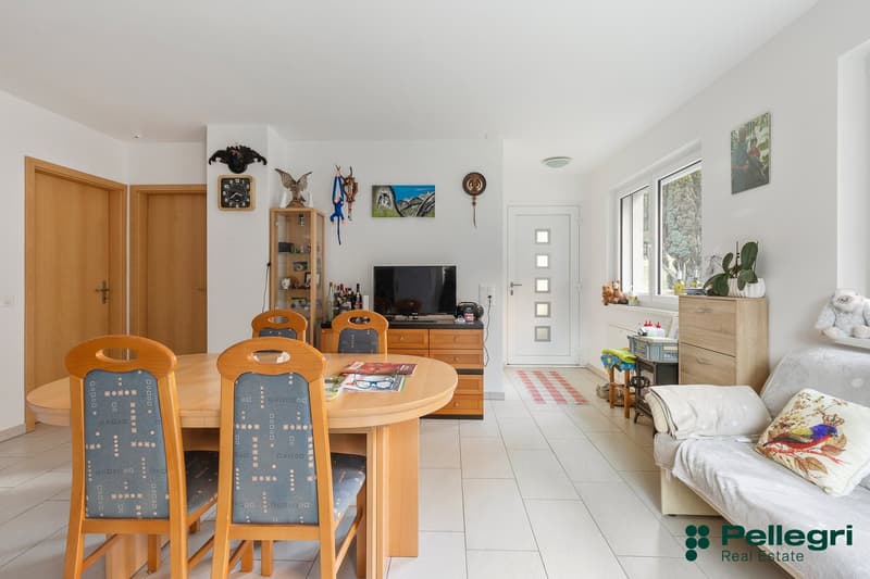 Modern living room with white tiles, wooden furniture, glass table, television, bookshelves, plants, and decorative items.