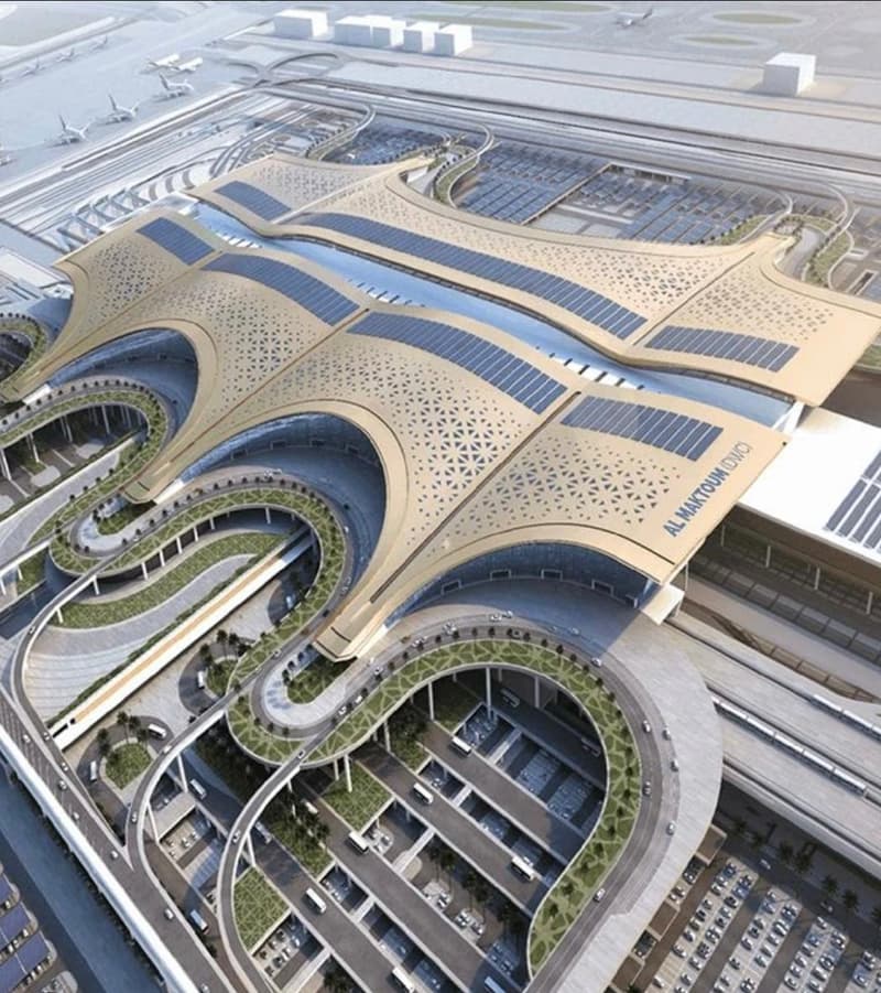 An aerial view of Al Maktoum International Airport, characterized by a distinctive spiral roof structure and extensive use of solar panels. The architectural design is modern with a blend of green spaces. There are numerous vehicles and buses visible in t