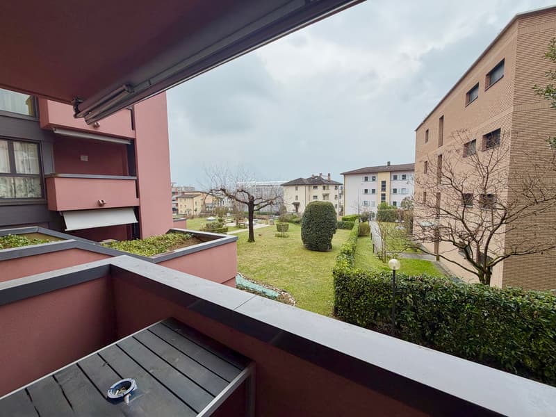 Balcony with a view of a garden and buildings, wooden floor, metal railing, green plants, grass, and trees