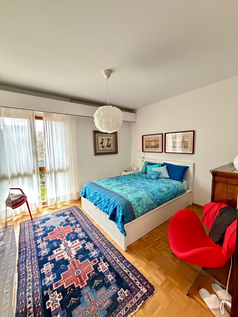 bedroom with bed, rug, two chairs, nightstand, three paintings, lamps, curtains