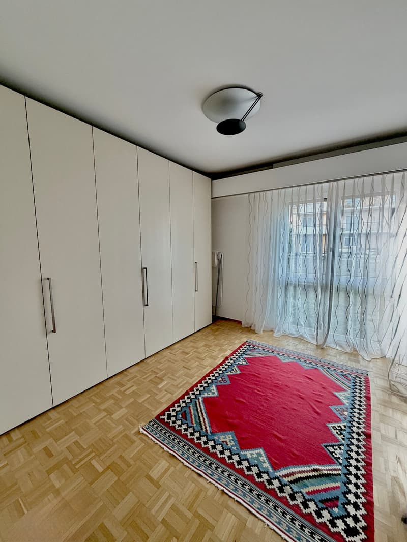Empty room with wooden floor, white walls, large windows with white curtains, built-in wardrobes, and a red rug