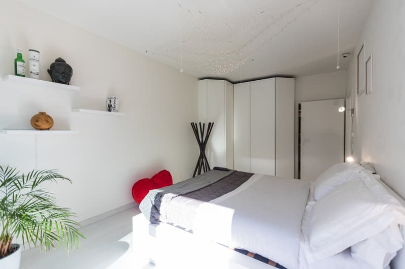 white walls, white bed, white shelves, white ceiling lamp, red bean bag, potted plant