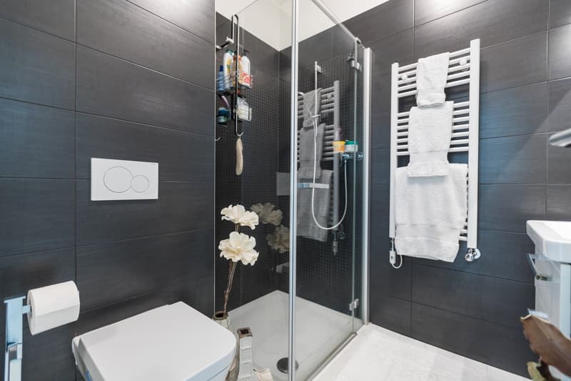 modern bathroom with white tiles, glass shower enclosure, white toilet, white sink, towel rack, flower vase, toilet paper holder
