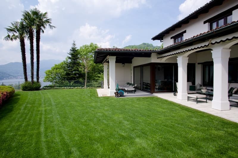 Two-story Mediterranean-style house with a tiled roof, surrounded by a well-manicured lawn and palm trees. The house has a large covered patio or terrace area with outdoor furniture, and the property offers a scenic view of the surrounding landscape.
