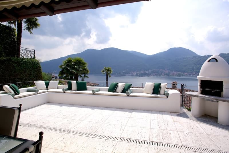 Spacious outdoor terrace with comfortable seating, overlooking a lake surrounded by mountains. The terrace has a tiled floor, a fireplace, and palm trees in the background.