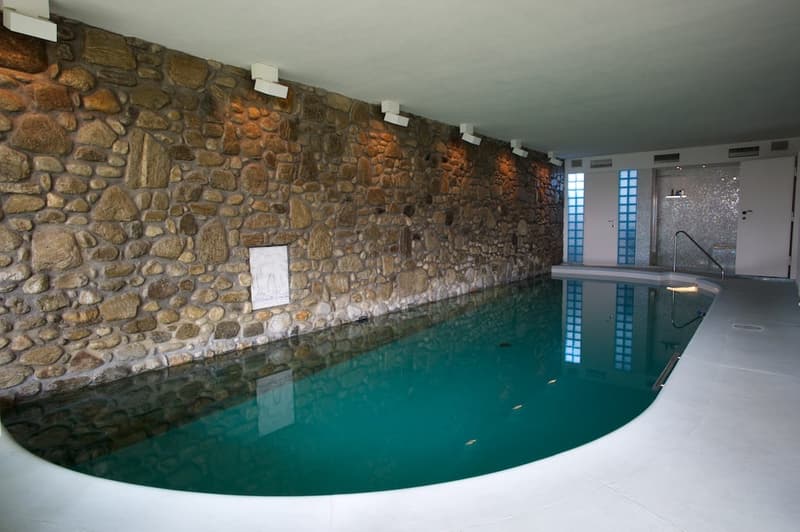 Large indoor swimming pool with a stone wall on one side, creating a rustic and cozy atmosphere. The pool has a clean, turquoise-colored water surface that reflects the lighting. The space appears to be part of a larger residential or hotel property.