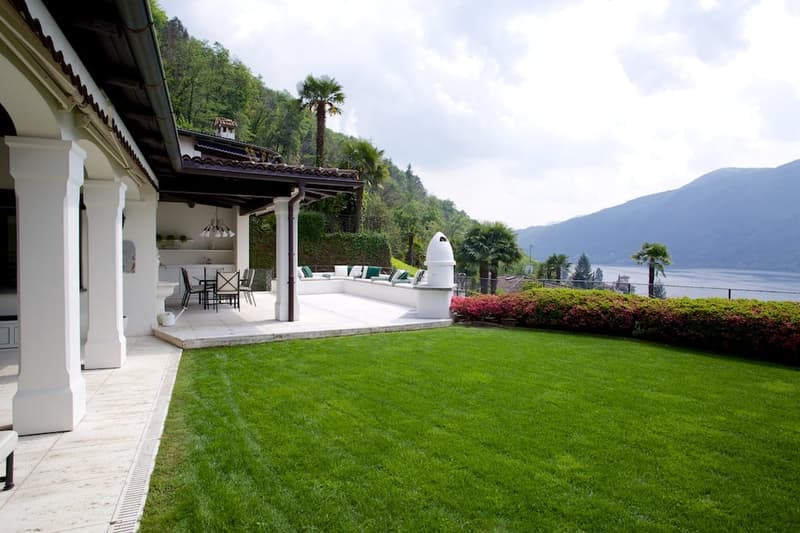 Two-story house with a covered patio, surrounded by a well-manicured lawn and lush greenery. The house has a Mediterranean-style architecture with a tiled roof and white walls. The patio features outdoor seating and a fireplace, offering a scenic view of