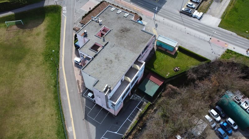 Aerial view of a 2-story house with a flat roof, car parked in front, grass, street, and parked cars