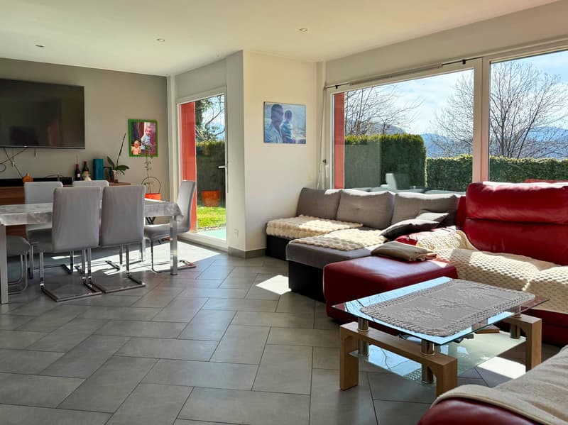 Spacious open-plan living area with large windows overlooking a balcony or terrace, hardwood floors, a red leather sofa, a dining table, and various decorative elements such as artwork and plants.