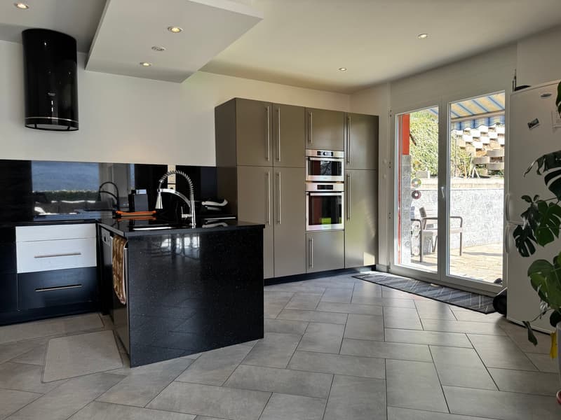 Modern kitchen with black granite countertops, gray cabinets, built-in appliances including a dishwasher and washing machine, and large windows providing natural light