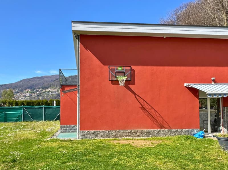 Two-story building with a red exterior, a basketball hoop on the side, and a grassy area in front. There appears to be a balcony or terrace visible on the upper level.
