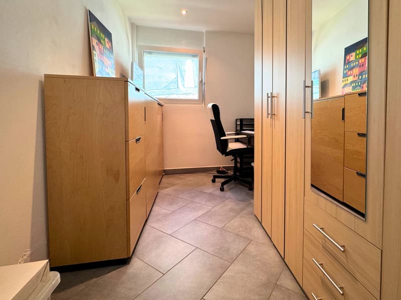 Small office space with wooden cabinets, a desk chair, and tiled flooring. The room has a window and appears to have a washing machine and dishwasher installed.