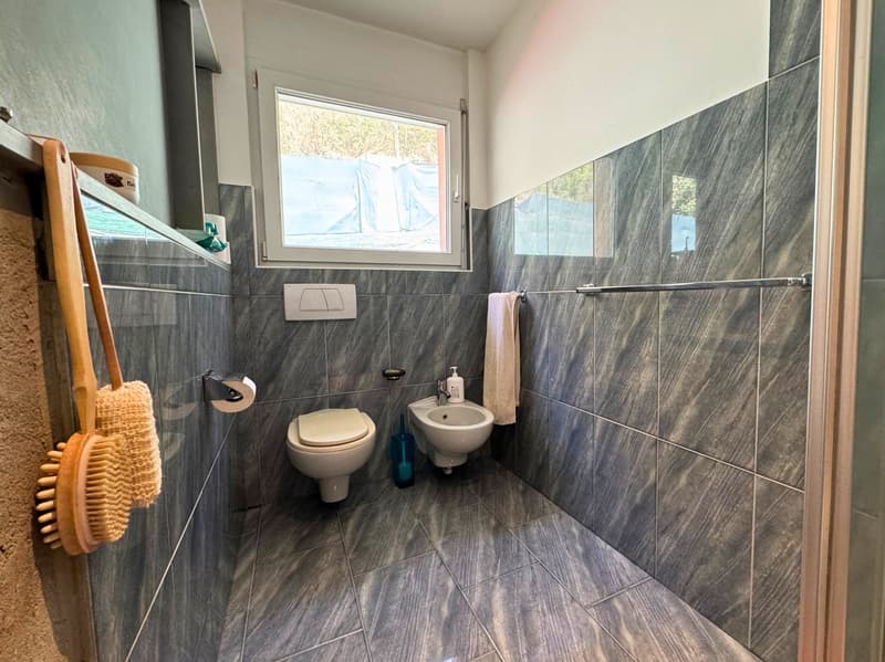 Bathroom with gray tile walls and floors, toilet, bidet, and a window overlooking the outdoors.