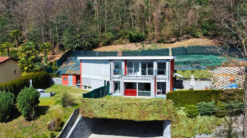 Two-story modern house with white and red exterior, large windows, surrounded by lush greenery and trees, with a balcony or terrace visible on the upper level.