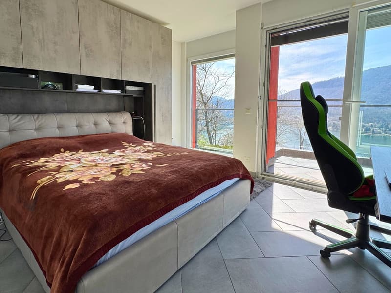 Spacious bedroom with large windows offering a scenic view of the mountains and lake. The room features a plush, floral-patterned comforter on the bed, built-in storage cabinets, and a comfortable-looking gaming chair by the window.