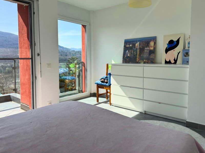 Spacious bedroom with large windows offering a scenic view, white walls, gray bedding, white dresser, and artwork on the wall