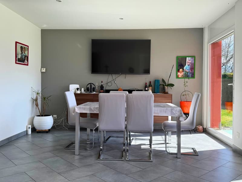 Spacious dining area with a large wooden dining table and white chairs, a mounted TV on the wall, and a sideboard with various decor items. The room has gray walls and a sliding glass door leading to a balcony or terrace.