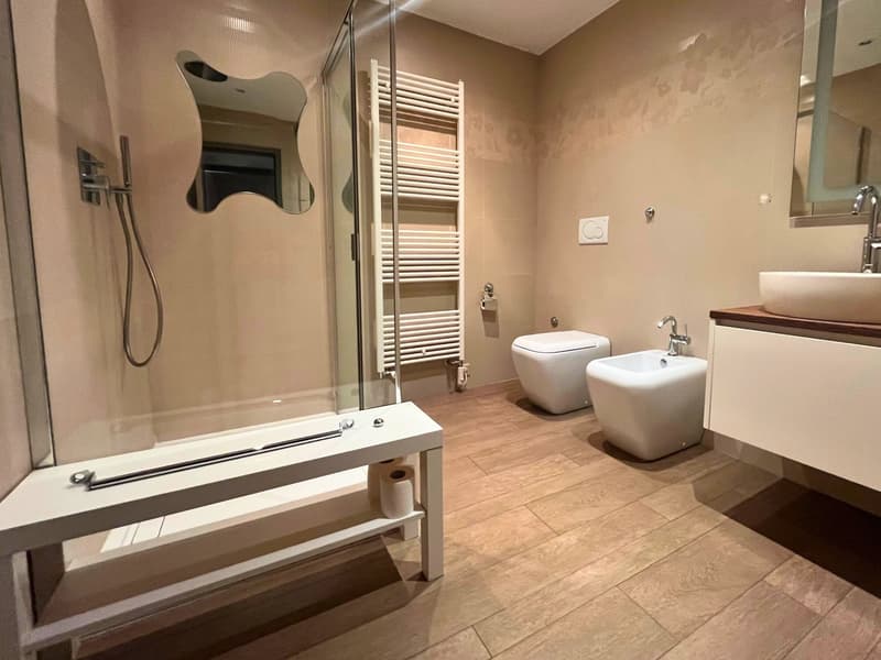 White shower area with shower head, white sink, white bidet, white toilet, white towel rack, white roll paper holder, white shelves, mirror, wooden floor
