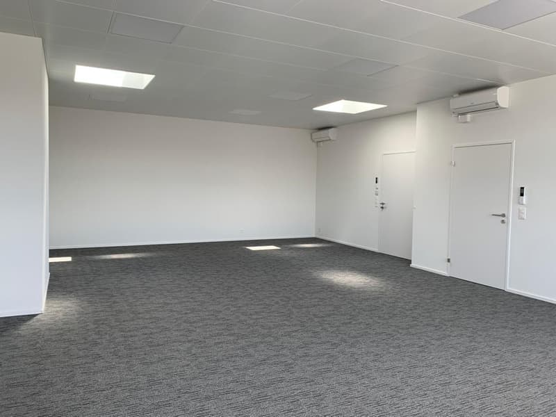 Large open office space with gray carpeted flooring, white walls, and recessed lighting. The room has a door and appears to be a commercial or business space.