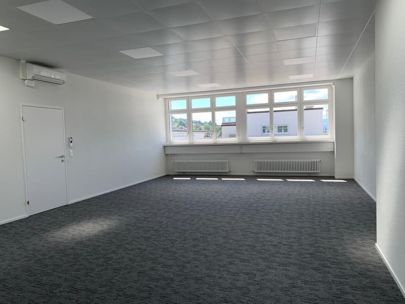 Large open room with white walls, gray carpeted floor, and multiple windows providing natural light. The room appears to be an office or commercial space with a clean and modern design.