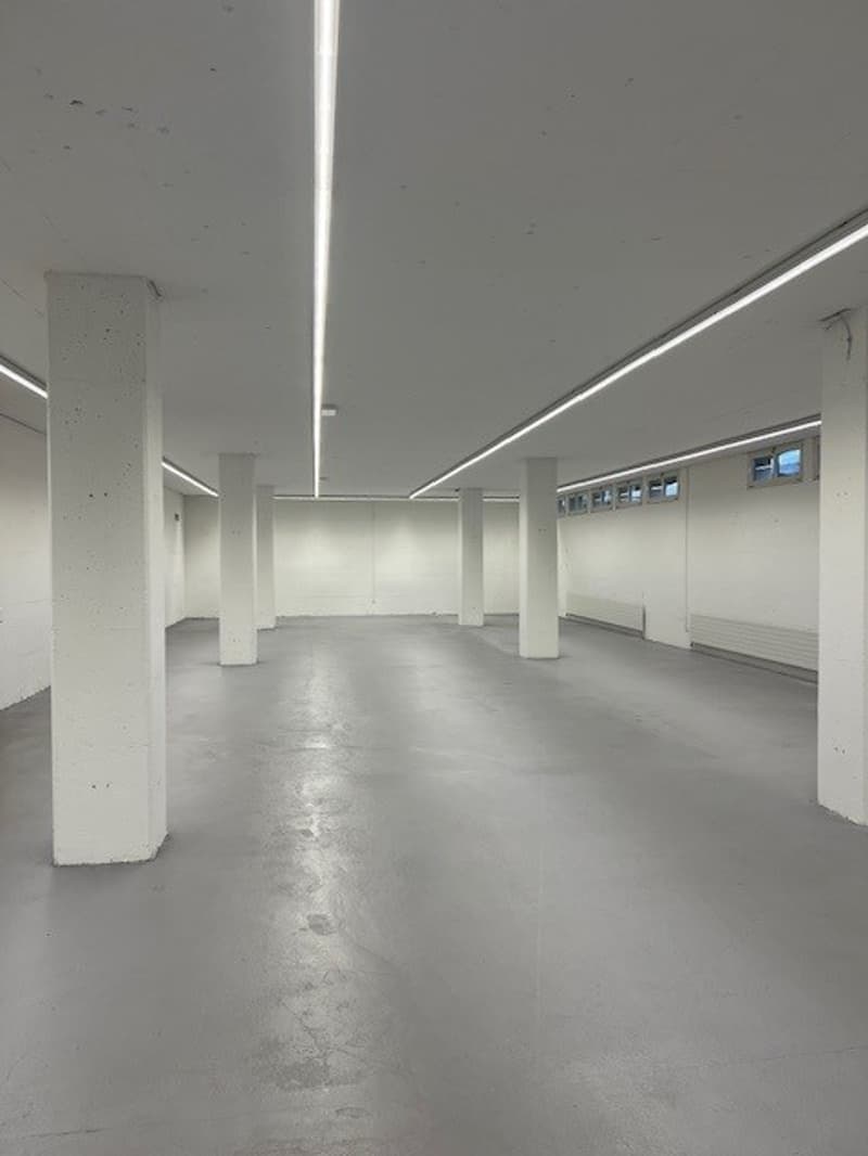 Large open space with concrete floor, white walls, and overhead lighting fixtures. The room appears to be an empty commercial or industrial space, possibly a warehouse or studio.