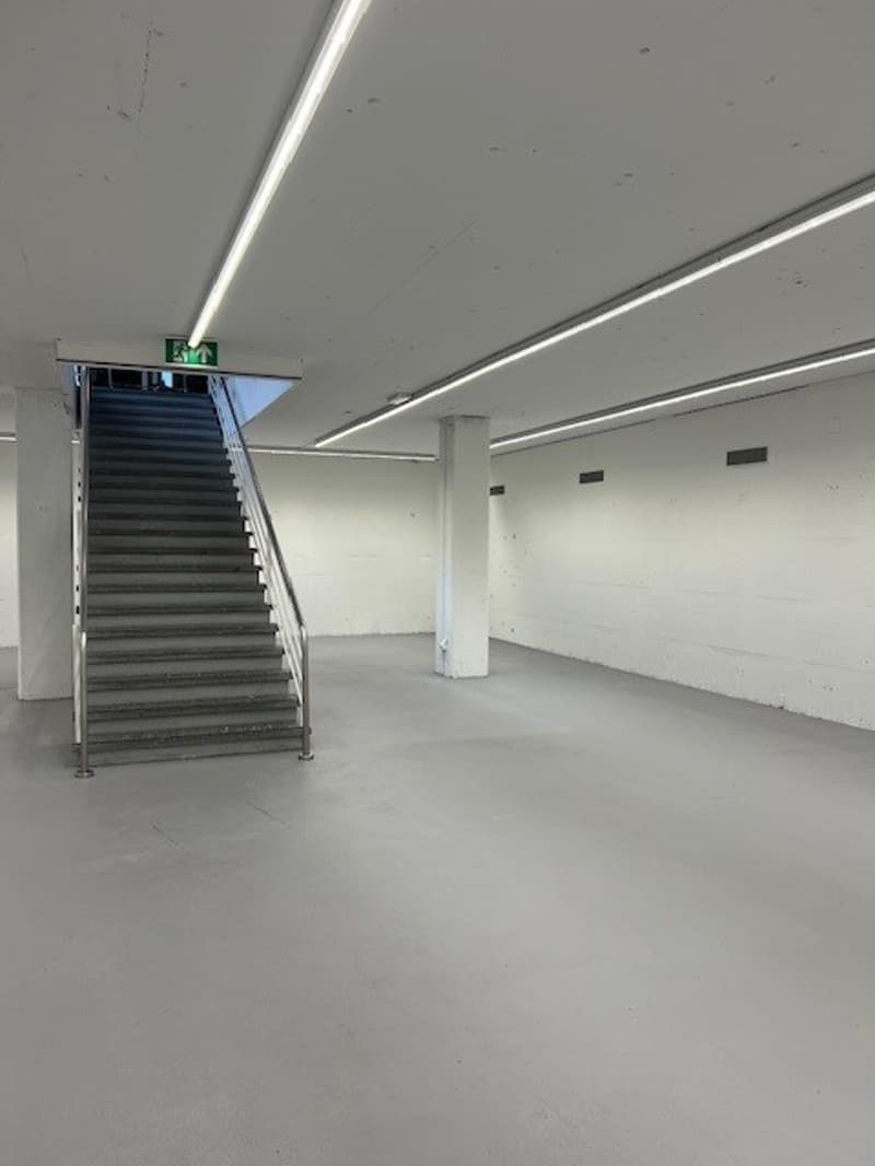 The image shows a large, open office space with a staircase leading up to a higher level. The space has a minimalist, modern design with white walls and ceiling, and long, linear lighting fixtures. The floor appears to be a smooth, gray surface, likely co