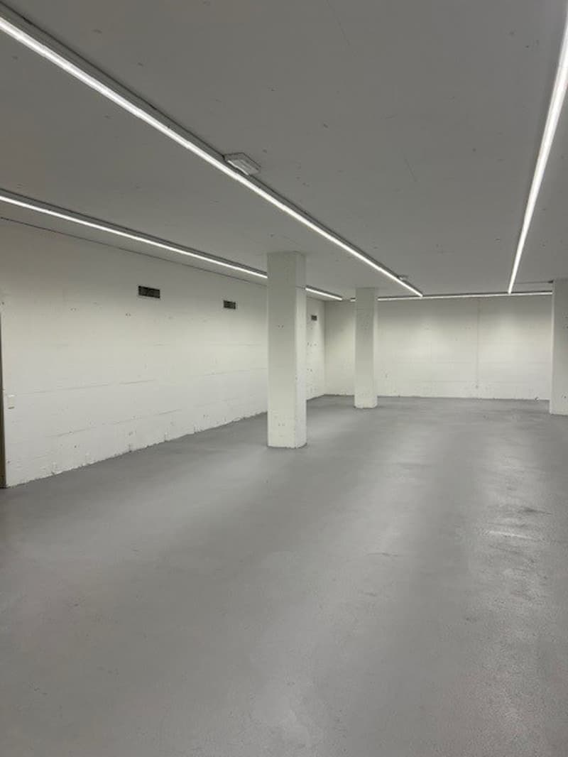 Large open space with white walls, concrete floor, and overhead lighting. The room appears to be an empty commercial or industrial space, possibly a warehouse or office area.