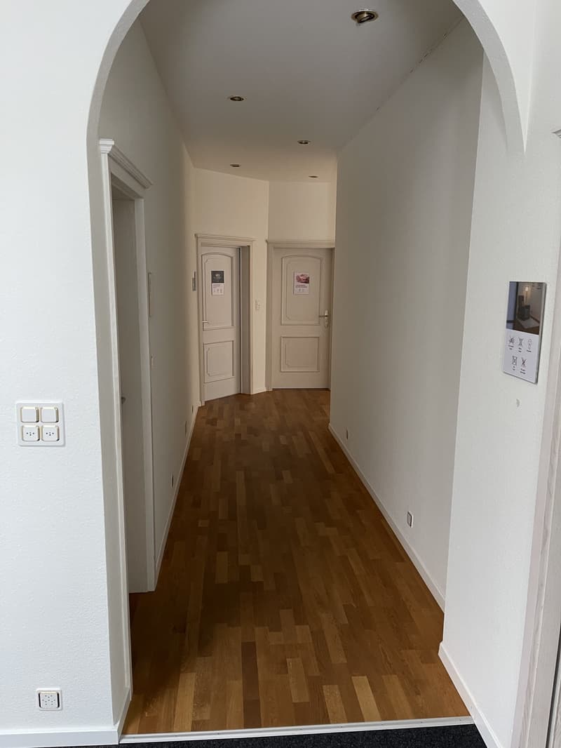 The image shows a hallway with white walls and a wooden floor. There are two doors on the left side of the hallway, which appear to be the entrances to separate rooms. The hallway is well-lit with recessed lighting in the ceiling.