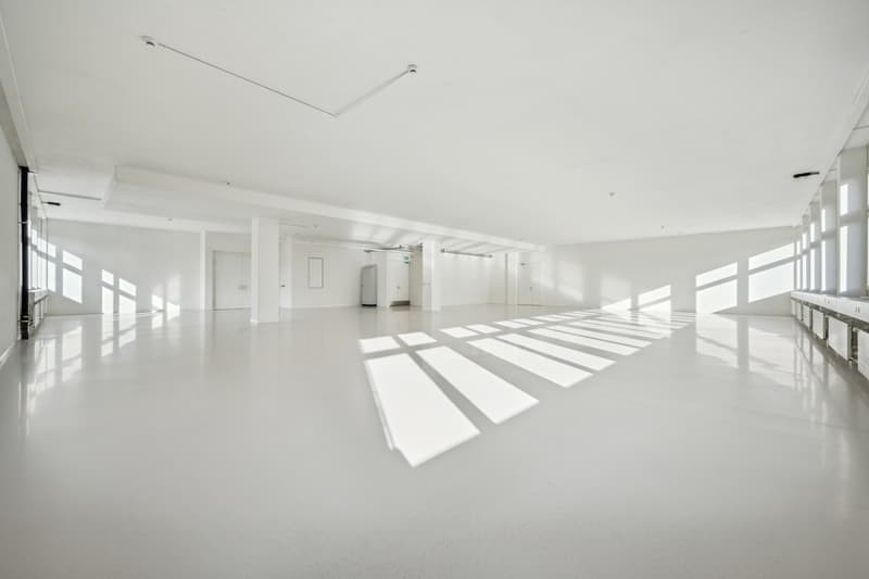 Large, open-plan office space with high ceilings, bright natural lighting from the windows, and a clean, minimalist design. The room appears to be empty, with a polished concrete floor and white walls.