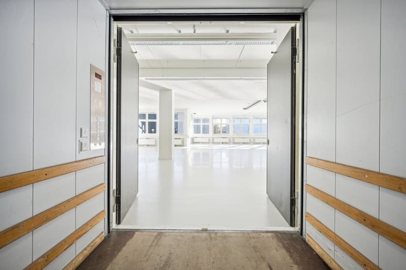 Large open office space with white walls, wooden accents, and large windows providing natural light. The space appears to be a commercial or industrial setting, potentially an office or studio.