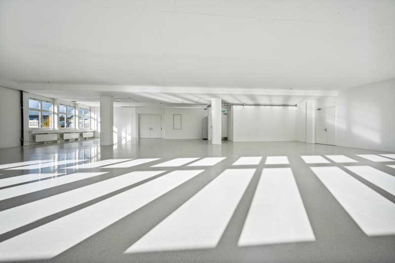 Large open-plan office space with white walls, high ceilings, and large windows allowing natural light to flood the room. The floor is a polished concrete surface with a geometric pattern. The space appears to be empty and ready for use.