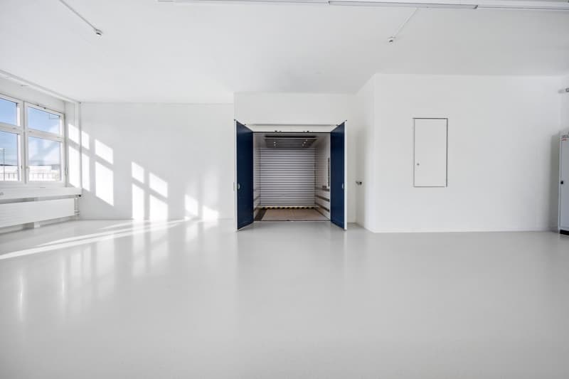 Large, open space with white walls and floors, large windows, and a garage door entrance in the center of the room.
