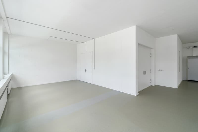 Spacious, minimalist room with white walls, gray flooring, and a door leading to another room. The room appears to be empty, with no furniture or decor visible.
