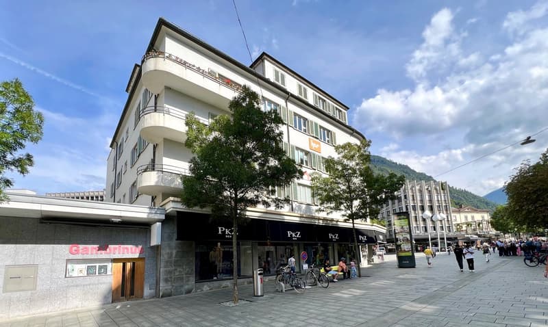 A multi-story commercial and residential building with balconies, located in a city center with a plaza and pedestrian area in front of it.