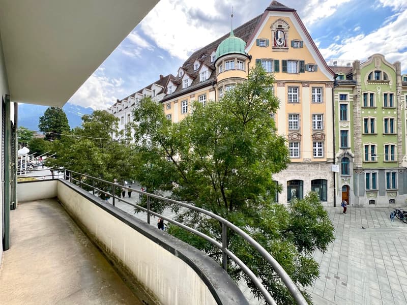 A multi-story historic building with a copper dome roof, surrounded by trees and greenery. The building has ornate architectural details and is situated on a plaza with pedestrians and bicycles visible.