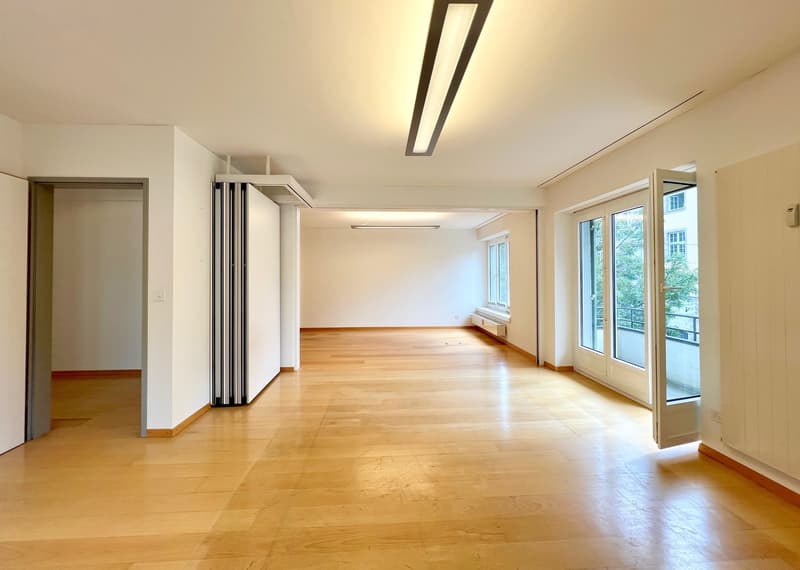 Spacious open-plan room with hardwood flooring, large windows providing natural light, and a sliding glass door leading to a balcony or terrace. The room has a minimalist design with white walls and a modern lighting fixture overhead.