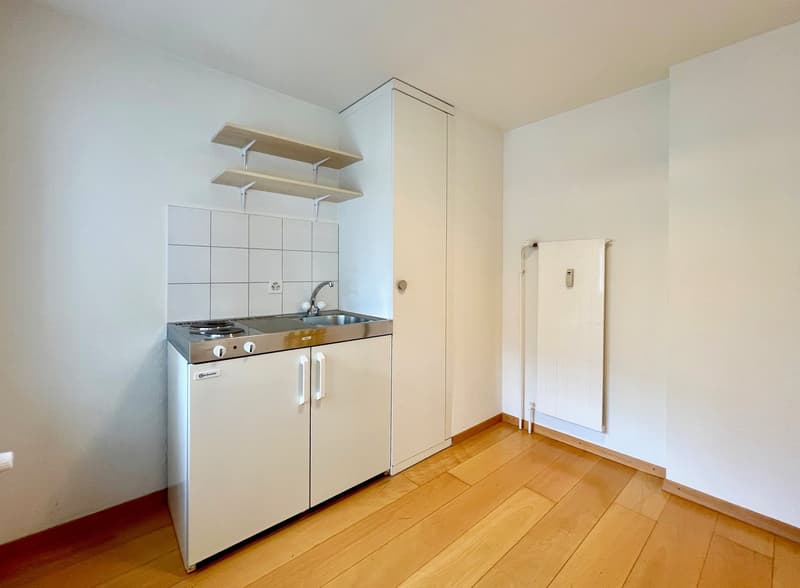 The image shows a small studio apartment with a kitchenette area. The kitchenette has a stove, oven, and sink, as well as some open shelving above it. The floor is made of hardwood, and the walls are painted white. There is a white door visible in the cor