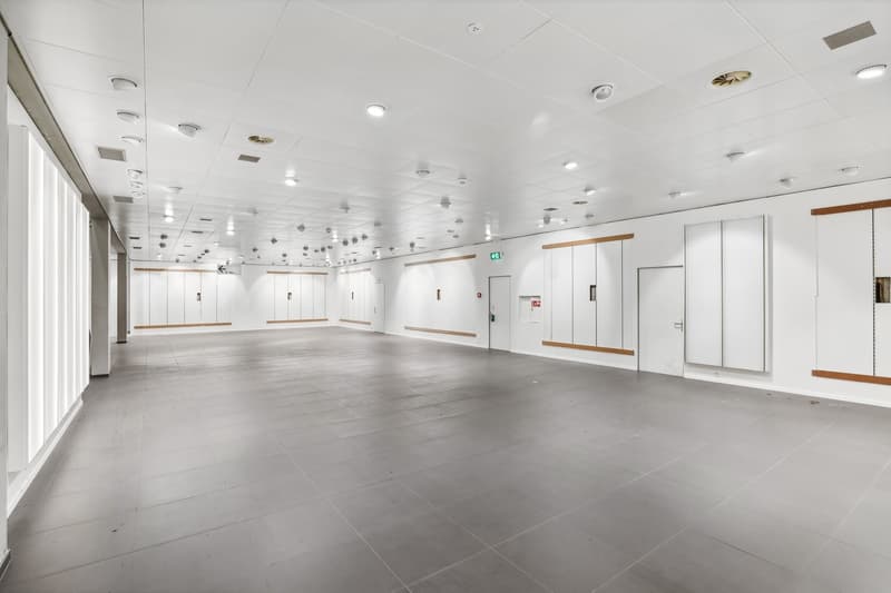 Large open space with white walls, gray tile flooring, recessed lighting, and several doors along the walls.
