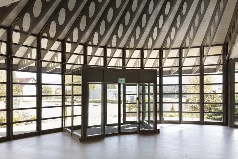 The image shows the entrance of a building with a curved glass ceiling and large windows. The entrance has glass doors and a sign indicating an exit. The interior is open and spacious, with a tiled floor and a view of the outdoor environment through the w