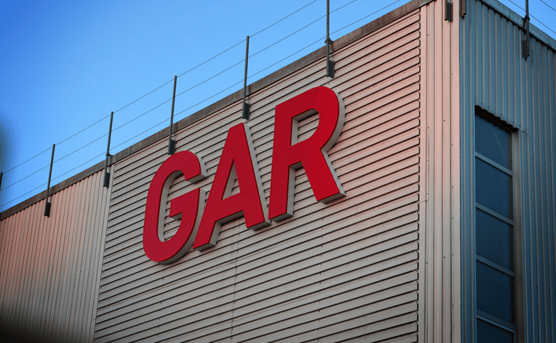 The image shows a large building with a prominent red 'GAR' sign on the side. The building appears to be made of wooden panels and has a blue sky in the background.