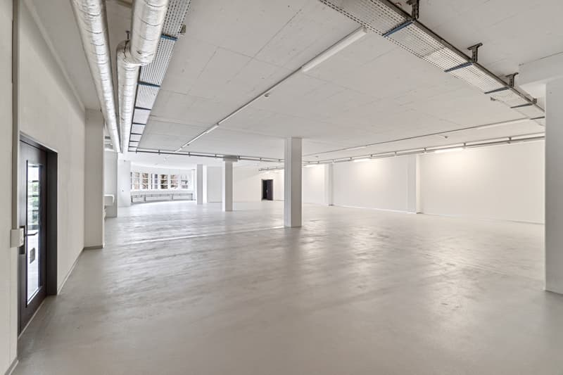 Large open-plan commercial space with high ceilings, exposed ductwork, and concrete flooring. The space appears to be a loft or warehouse-style office, with multiple windows and a door leading to another area. The space is currently empty and unfinished,