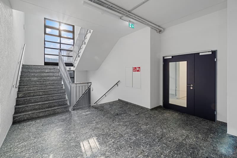 The image shows the interior of a building, with a staircase leading up to the upper floors. The walls are painted in a light color, and the floor is covered in gray granite tiles. There is a fire exit sign on the wall, and a black door leading to another