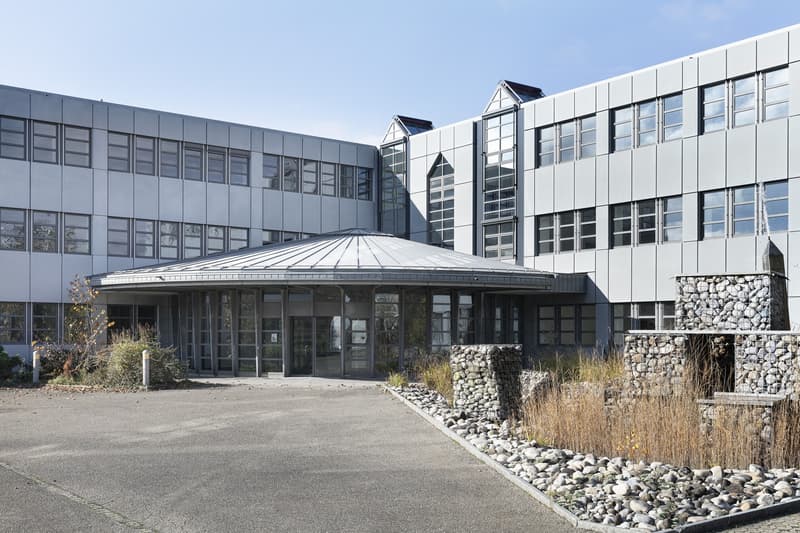 Large modern commercial building with a glass and metal facade, a domed roof structure, and a paved parking area in the foreground. The building has multiple floors and large windows. There is also a gabion wall feature with stones near the entrance.