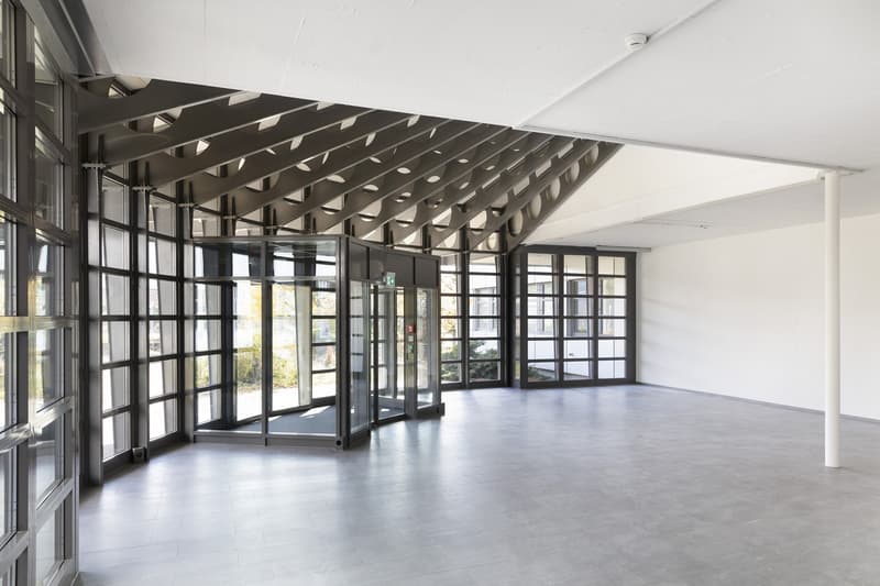Large open-plan office space with high ceilings, glass walls, and a modern geometric ceiling design. The space has a minimalist and industrial aesthetic, with concrete floors and a bright, airy atmosphere.