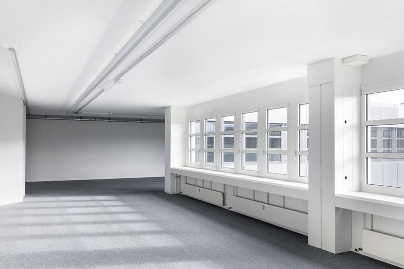 Large open office space with high ceilings, white walls, and large windows providing ample natural light. The room has a gray carpeted floor and appears to be set up for office use with potential for various workstations or meeting areas.