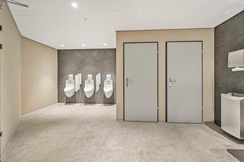 The image shows a commercial bathroom with multiple white urinals mounted on a gray tiled wall. There are two gray doors, likely leading to individual stalls or restrooms. The floor is covered in a light-colored tile or concrete. The overall design appear