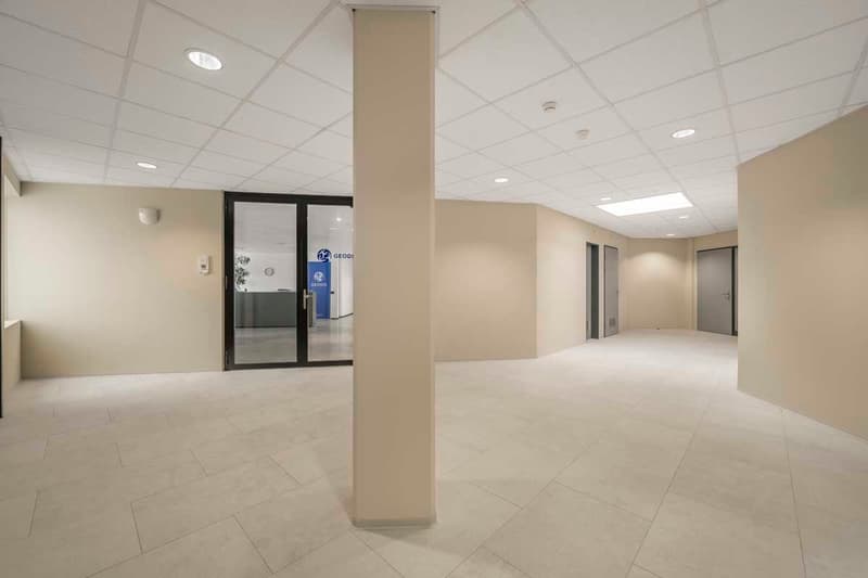 The image shows a large, open office space with a tiled floor, recessed lighting, and a glass-paneled door leading to another room. The space appears to be a commercial or office environment, with a clean and modern design.